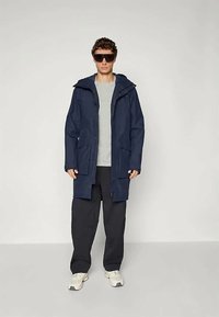 Navy waterproof parka with a hood, front zipper, and two side pockets, styled over a grey t-shirt and dark pants, paired with sunglasses.