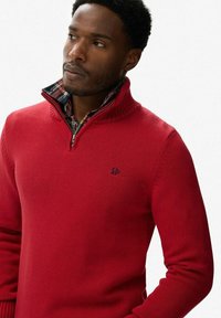Suéter rojo de punto con cuello con cremallera, que presenta un forro interior a cuadros y un pequeño logo en el pecho. Tejido texturizado y diseño ajustado.