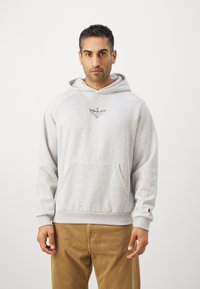 Felpa grigia con cappuccio, tasca a marsupio, dettaglio del logo sul petto, polsini e vita a coste, indossata con pantaloni di velluto a coste.
