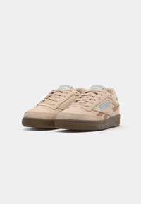 Sneakers in suede beige chiaro con suole in gomma marrone, dettagli blu e arancione e un design testurizzato. Presentano un sistema di chiusura con lacci e un particolare con il logo.