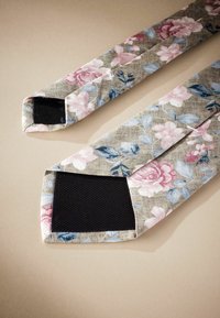 Blumenmusterte Stoffriemen in Rosa und Blau auf einem beigen Hintergrund, mit schwarzen Netzakzenten an den Enden. Rechteckige Form, strukturierte Oberfläche.