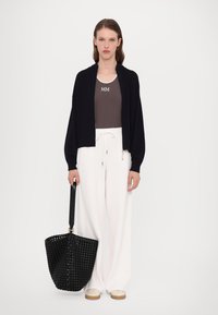 Svart strikket cardigan over en grå tanktop med "MM" tekst, kombinert med hvite vide bukser og en svart vevd toteveske med en solid stropp.