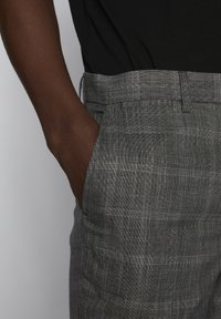 Šedé vzorované kalhoty z tkané látky, s plochým předním dílem a bočními kapsami, detail na pas a ruku v kapse.