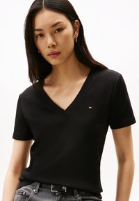 Fiatal nő hosszú, sötét hajjal, fekete V-nyakú pólóban és szürke farmerben, előre nézve, semleges arckifejezéssel.