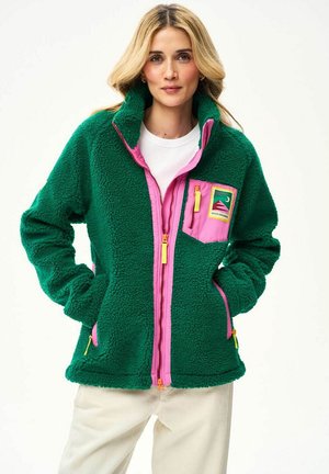Kobieta w zielonym polarowym swetrze z różowym zamkiem błyskawicznym i kieszenią, stojąca z rękami w kieszeniach na jednolitym tle.
