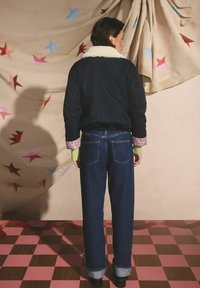 Jachetă bomber bleumarin închis cu guler alb pufos, detalii florale la manșete; combinată cu blugi albaștri cu talie înaltă și pantofi negri, pe un podea în carouri.