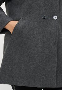 Γκρίζο παλτό από μείγμα μαλλιού με ετικέτα κολάρο, διπλή κουμπωμένη σχεδίαση, δύο μαύρα κουμπιά και πλαινές τσέπες. Υφή επιφάνειας, μεσαίου μήκους.