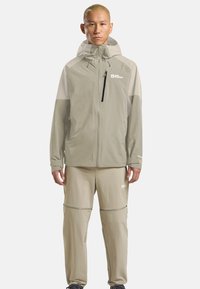 Uomo che indossa un giubbotto beige a due toni con cappuccio e pantaloni abbinati con zip, in piedi contro uno sfondo neutro.