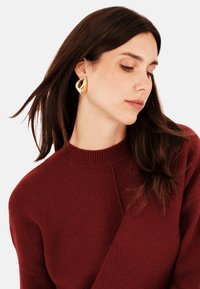 Zlaté, kulaté visací náušnice s texturovaným povrchem, které nosí osoba s dlouhými tmavými vlasy v burgundském svetru.