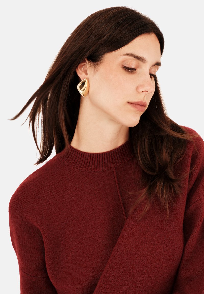 Zlaté, kulaté visací náušnice s texturovaným povrchem, které nosí osoba s dlouhými tmavými vlasy v burgundském svetru.