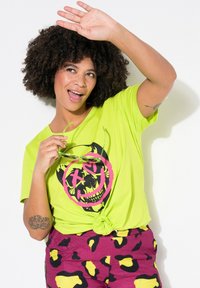 T-shirt vert néon avec un graphique de crâne noir et rose, noué à la taille, associé à un short violet avec un motif audacieux jaune et noir.