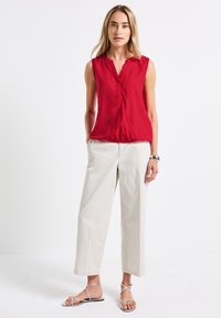 Chemisier sans manches rouge avec un col en V et un pli devant, associé à un pantalon beige à jambes larges et des sandales à brides argentées.