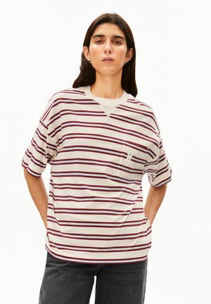 Femme aux longs cheveux foncés, portant un t-shirt blanc à manches courtes avec des rayures rouges horizontales et un pantalon noir, debout les mains dans les poches.