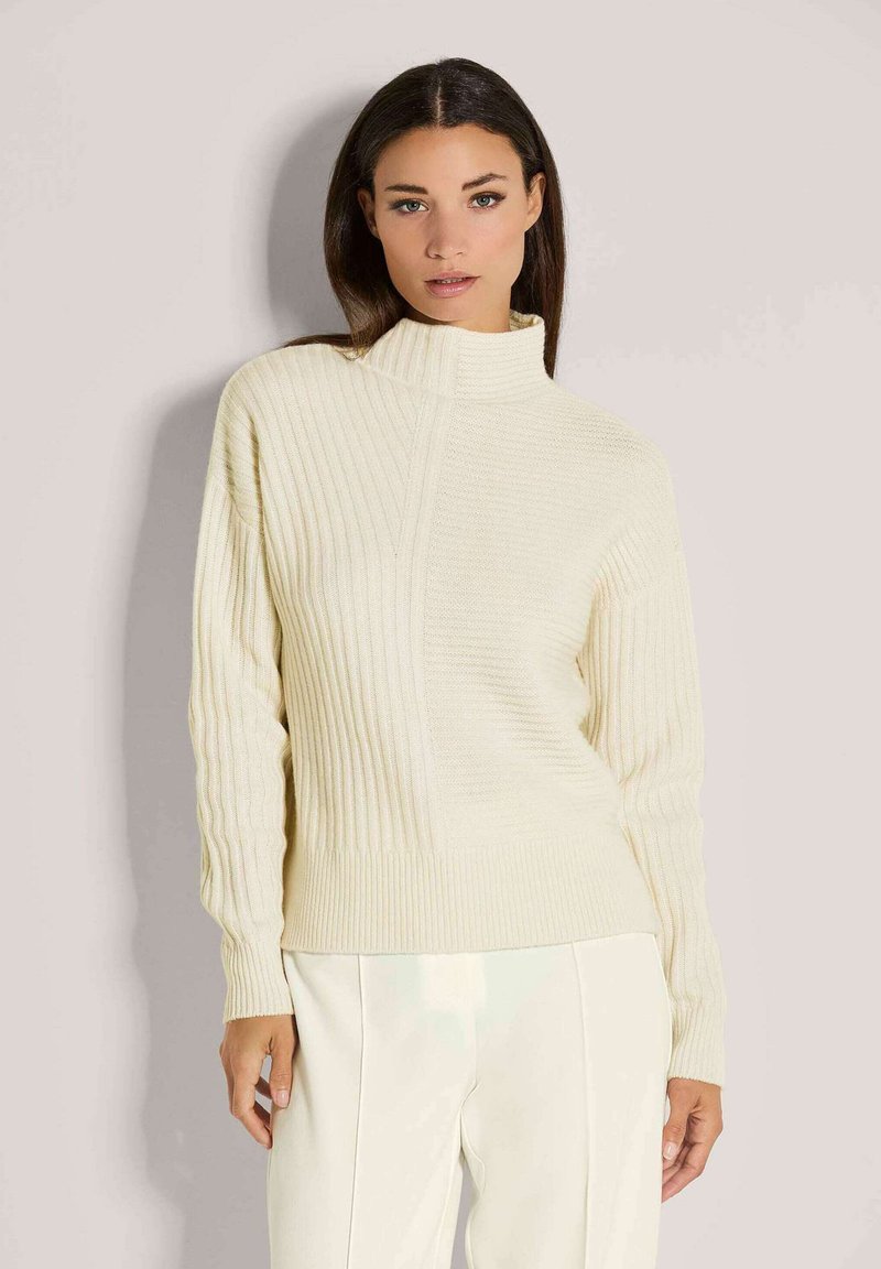 Vrouw met lang donker haar, gekleed in een crèmekleurige ribgebreide trui met hoge nek en bijpassende crèmekleurige broek, staande tegen een effen achtergrond.