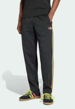 Osoba ubrana w czarne spodnie dresowe z różowo-pomarańczowo-żółtymi paskami po bokach oraz pasujące czarne trampki z neonowożółtymi sznurowadłami.