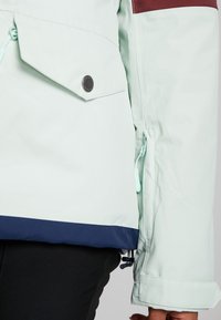 Jasno miętowa zielona kurtka z bordowym akcentem na ramieniu, detalami z guzików, boczną kieszenią oraz granatowym obszyciem na dole. Wytrzymała tekstura tkaniny.