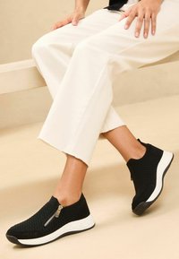 Sneakers in maglia nera con tomaia texturizzata, suola bianca e dettaglio in zip argentato. Abbinati a pantaloni bianchi cropped.