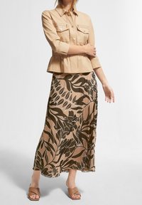 Veste courte beige avec deux poches devant, associée à une longue jupe à motifs floraux dans des tons de marron et crème, portée avec des sandales marron.