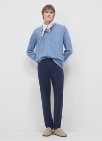 Camicia azzurro chiaro con una texture morbida e sciarpa beige, abbinata a pantaloni plissettati blu scuro e scarpe slip-on beige.