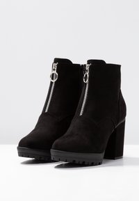 Botines de ante negros hasta el tobillo con tacón cuadrado, que cuentan con un cierre frontal con tirador plateado. Suela de goma texturizada para mayor agarre.