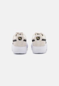 Baskets en suède beige avec une semelle blanche, présentant des rayures noires et un accent de logo noir sur le talon. Texture douce, design décontracté.