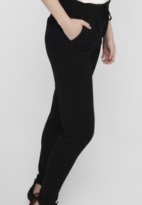 Vrouw in zwarte strakke broek met een trekkoord in de taille en zakken, staand met één hand in de zak en gekleed in zwarte hakken.