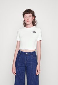 Camiseta blanca de corte corto con mangas cortas y un logo negro en el lado izquierdo del pecho. Combinada con pantalones de mezclilla azules de cintura alta.
