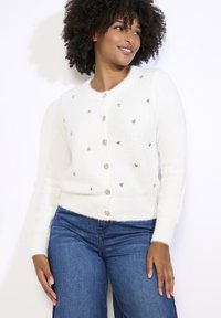 Bílý, načechraný kardigan s kulatými, stříbrnými ozdobami. Má kulatý výstřih, dlouhé rukávy a zapínání na knoflíky vpředu.