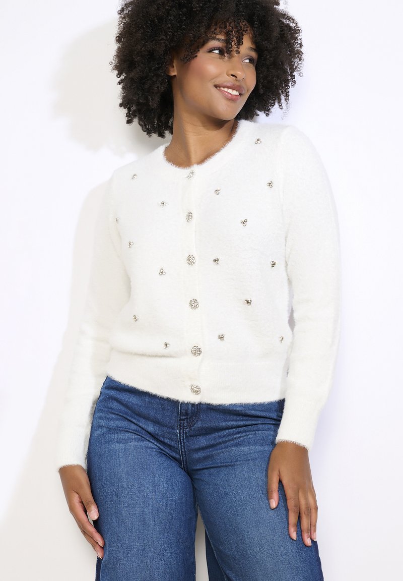 Bílý, načechraný kardigan s kulatými, stříbrnými ozdobami. Má kulatý výstřih, dlouhé rukávy a zapínání na knoflíky vpředu.