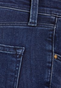 Tmavě modré denimové džíny s pětistopým designem, viditelným zlatým kovovým knoflíkem a prošitím podél pásu a kapes.