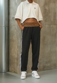 Kortärmad vit polo med krage, svarta sweatpants med vita ränder på sidorna, och vita sneakers med orangea accenter.