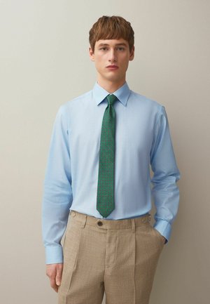 Young man wearing a light blue dress shirt, green patterned tie, and beige trousers, standing with one hand in pocket against plain background.