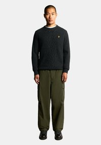 Suéter de punto grueso gris oscuro con cuello redondo, combinado con pantalones cargo color oliva y botas negras con cordones. Detalle de logo amarillo en el suéter.