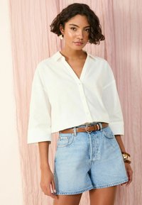 Camicia bianca con maniche a tre quarti, pantaloni corti in denim blu, cintura marrone e bracciali a fascetta. Tessuto morbido; abbigliamento informale.