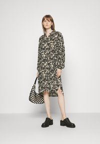 Vestido floral em preto e bege com mangas longas, bainha dividida, combinado com mules de salto bloco pretos e uma bolsa de rede preta.