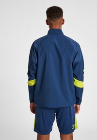 Marinblå sportjacka med hög krage, med fluorescerande gula accentremsor på ärmarna. Slät, lättviktsdesign.