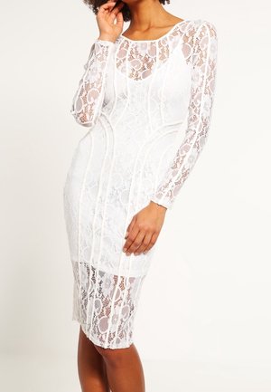 Femme portant une robe blanche en dentelle à manches longues, longueur genou, avec des motifs floraux et des coutures ajustées, posant avec une main près de son visage.