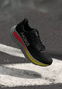 Shoe atlético preto com um cabedal texturizado, apresentando um logotipo branco e uma sola em várias cores com gradientes de amarelo, laranja e vermelho.