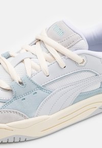 Bílé a světle modré tenisky s materiály z měkké síťoviny a semiše, plochými tkaničkami, strukturovanými panely a gumovou podrážkou s viditelným vzorem.