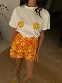 White T-shirt with orange slice appliqués, paired with orange shorts featuring a citrus pattern. Both garments have relaxed fits and soft textures.