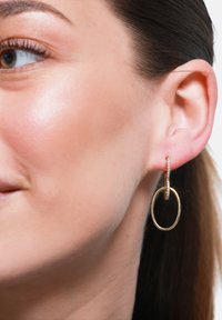 Orecchini a cerchio in oro con un design circolare più piccolo che presenta piccoli cristalli trasparenti. Gli orecchini sono attaccati al lobo dell'orecchio.