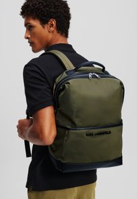 Rucsac verde măsliniu din nylon neted, cu un compartiment principal cu fermoar, accente negre și un logo pe față. Bretele ajustabile.