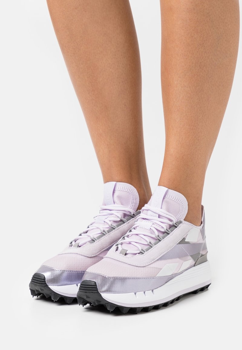 Light purple athletic shoes with a mix of matte and shiny textures, featuring a layered design, prominent laces, and a black rubber sole.
