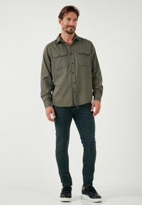 Olijfgroene blouse met knoopsluiting en twee voorzakken, gecombineerd met donkere jeans en zwarte instapschoenen, gedragen door een model tegen een effen achtergrond.