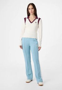 Hvid langærmet top med dyb V-hals og vinrøde accenter, kombineret med lyseblå bukser med sidelommer og et lille logo.