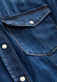 Detaliu al unei cămăși din denim de culoare albastru închis, cu un buzunar ascuțit, nasturi albi cu capse, cusături contrastante de culoare portocalie și o textură netedă.