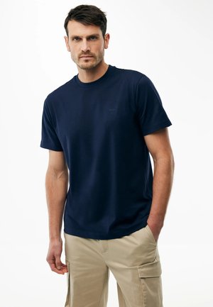 Mężczyzna o krótkich ciemnych włosach, ubrany w granatowy T-shirt i beżowe spodnie bojówki, stojący z jedną ręką w kieszeni na białym tle.