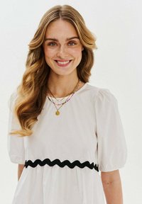 Biele šaty s nafúknutými rukávmi, s čiernym vlnitým pásom. Modelka má na sebe viacero zlatých náhrdelníkov a dlhé, vlnité vlasy.