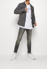 Hombre vestido con una camiseta gráfica blanca, una camisa abotonada gris oscuro, unos vaqueros desgastados grises arremangados y tenis blancos, apoyado contra un fondo sencillo.