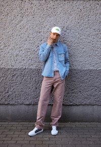 Světle modrá denimová košile, šedé tričko, růžové cargo kalhoty, bílé tenisky s květinovými motivy a bílá čepice se zeleným logem na pozadí texturované stěny.